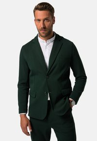 Blazer su misura verde scuro in tessuto liscio, con chiusura a due bottoni, rever a taglio e tasche laterali. Indossato sopra una camicia bianca.