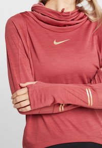 Red athletic long-sleeve top with a hood, featuring a gold Nike logo, thumbholes, and textured fabric with gold stripe accents on sleeves.
