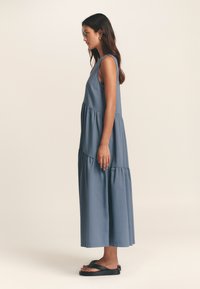 Maksikodas mėlynos spalvos, pagamintas iš minkštos medžiagos. Be rankovių, su sluoksniuotu stiliumi ir lengvai krentančia sijono dalimi, derinamas su juodomis sandalais.