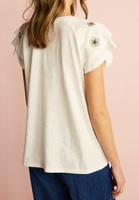 Camiseta de algodón blanca con mangas con volantes, presenta bordado floral en los hombros, corte relajado y una textura suave.