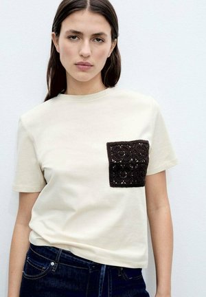 Crème-kleurig t-shirt met een getextureerde zwarte gehaakte zak, ronde halslijn, korte mouwen en een relaxte pasvorm.