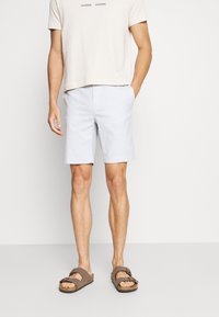 Muž v modrých šortkách, béžovém tričku s nápisem "SAMSOE SAMSOE" a hnědých sandálech s otevřenou špičkou, stojící s jednou rukou v kapse.