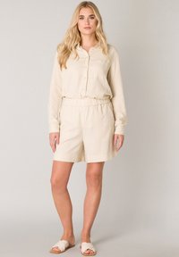 Beige linnen blouse met lange mouwen, knoopsluiting en borstzak, gecombineerd met bijpassende shorts met elastische taille. Platte sandalen maken de look af.