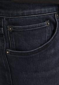 Pantalones vaqueros de mezclilla azul oscuro con un diseño clásico de cinco bolsillos, con herrajes en tono cobre y una sutil textura visible en la tela.