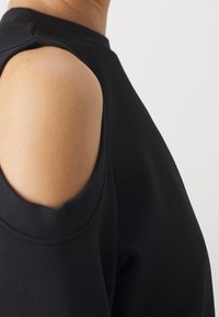 Primer plano de una persona que lleva una blusa negra de hombros descubiertos con el hombro izquierdo expuesto y un escote redondo, contra un fondo blanco.