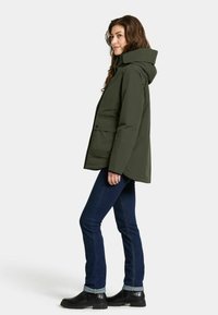 Donkergroene parka met capuchon, met een rits- en knoopsluiting, twee voorzakken en een ontspannen pasvorm. G gedragen met donkere jeans en laarzen.