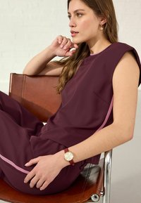 Maroon sleeveless top with lavender side detail, paired with matching pants. Wearing a burgundy watch with a gold face, seated on a leather chair.