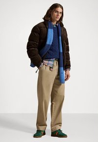 Brązowa kurtka puchowa, niebieski sweter w warkocze, koszula w kratę, beżowe spodnie, niebieski szalik w paski, zielone buty sportowe z pomarańczowymi akcentami.