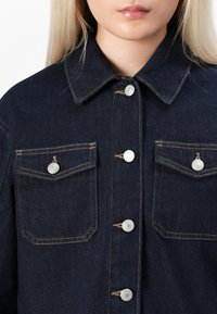 Femme portant une veste en denim bleu foncé avec des boutons argentés et deux poches poitrine boutonnées, montrée du cou au milieu du torse.