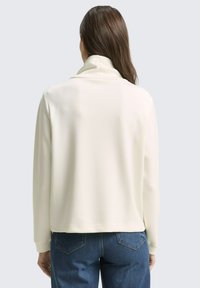 Crème-kleurige, lichtgewicht jas met een hoge kraag en ribbeltextuur; relaxte pasvorm; gedragen over blauwe jeans.