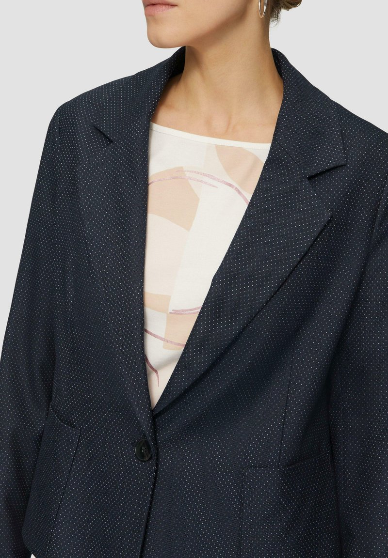 Donkerblauwe blazer met een gestippeld patroon, voorzien van een scherpe kraag, twee zijzakken en een enkele knoopsluiting.