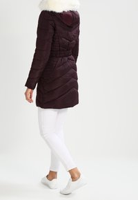 Burgundy pufferjacka med chevron-quiltat mönster, elastiskt midjeband och avtagbar vit faux fur-huva. Bärs över vita tighta byxor och sneakers.