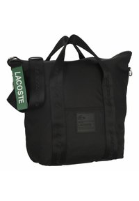 Sac fourre-tout Lacoste noir avec des poignées doubles, une bandoulière amovible et une étiquette verte affichant le nom de la marque Lacoste.