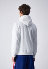 Sudadera blanca con capucha texturizada, material de forro polar suave, puños ajustados y un ajuste relajado, llevada sobre pantalones cortos deportivos azules.