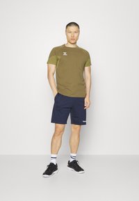 T-shirt vert olive avec un short bleu marine, ornée d'un logo sur la poitrine et sur le côté. Associée à des chaussures de sport noires et des chaussettes rayées.