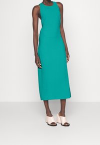 Tyrkysové bezrukávové midi šaty s přiléhavým střihem a bočním rozparkem, spárované s bledě růžovými otevřenými mules na hrubém podpatku.