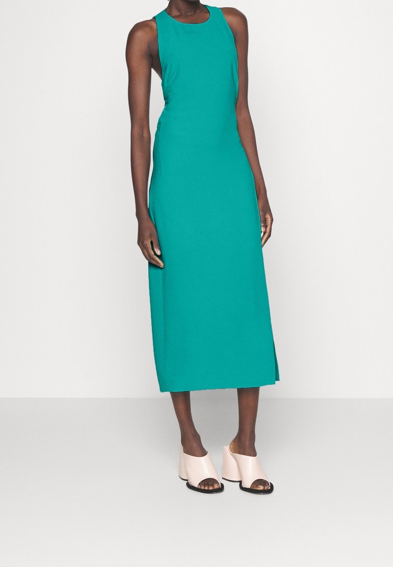 Tyrkysové bezrukávové midi šaty s přiléhavým střihem a bočním rozparkem, spárované s bledě růžovými otevřenými mules na hrubém podpatku.