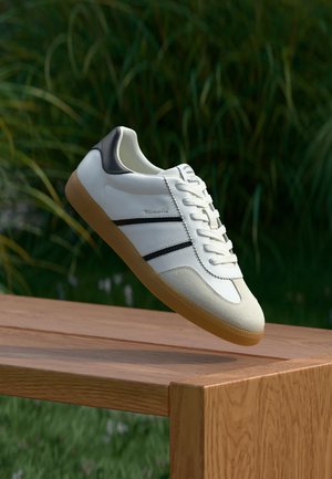 Weißer Sneaker mit schwarzen Streifen und Gummisohle, der über einer Holzbank im Freien schwebt, mit grünem Laub im Hintergrund.