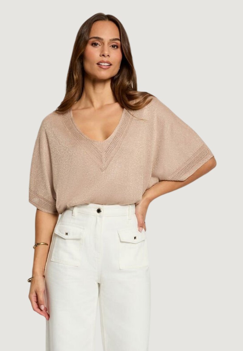 Woman wearing a beige short-sleeve V-neck sweater and high-waisted white pants with front flap pockets, posing with one hand on hip.