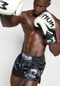 Pantalones cortos de boxeo de satén negro con una cintura elástica, logo de Venum en blanco y guantes de boxeo coordinados en negro y blanco. Se destaca una musculatura prominente.