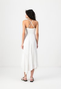 White maxi dress with a smocked bodice, adjustable crisscross straps, and a tiered skirt. Paired with light-colored sandals.