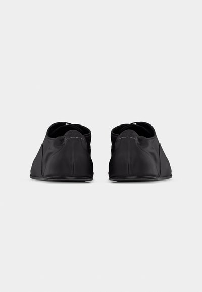 Ζευγάρι μαύρα παπούτσια χωρίς κορδόνια, ορατά από πίσω, με ελαστικές λεπτομέρειες και λεία, ματ επιφάνεια, πάνω σε απλό ανοιχτό φόντο.