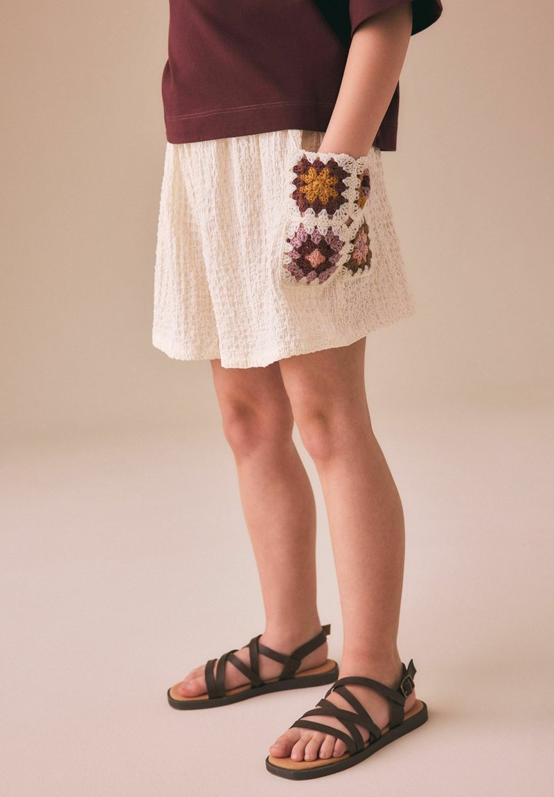 Kind trägt strukturierte weiße Shorts, burgunderfarbenes Oberteil, schwarze Riemchensandalen und eine farbenfrohe gehäkelte Tasche auf der rechten Seite.