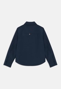 Mørkeblå skjorte med lange ermer, spiss krage, knappeermer og en liten logo-patch i rødt, hvitt og blått øverst på ryggen. Glatt tekstur.