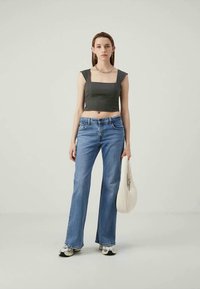 Mlada ženska stoji v sproščenem oblačilu: črn cropped top, modre kavbojke, športni copati, srebrna ogrlica in uhani s kavljem, drži belo torbico.