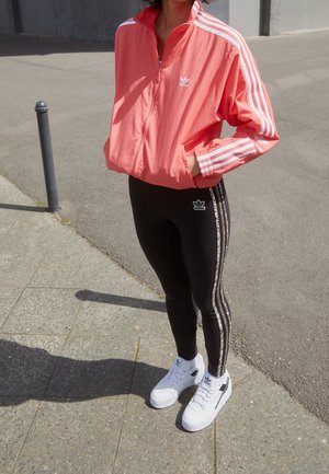 Person iført koralfarvet Adidas jakke og sorte Adidas leggings med leopardstribede detaljer, stående på en brolagt gade i hvide sneakers.
