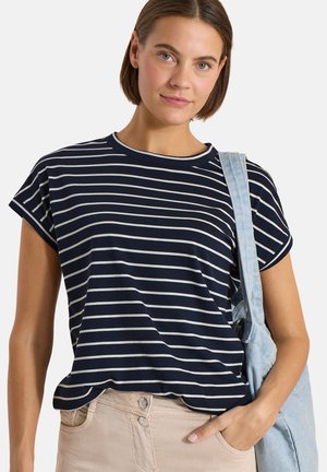 Femme portant une chemise à manches courtes rayée bleu marine et blanche, pantalon beige, et portant un sac à bandoulière bleu clair, faisant face à l'avant avec une main dans la poche.