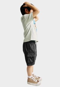 T-shirt gris clair avec un design graphique, shorts cargo noirs et baskets montantes beiges avec des lacets blancs et des accents. L'enfant se tient debout, les mains sur la tête.