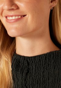 Gros plan sur une personne portant un haut noir texturé avec un design froncé et une petite boucle d'oreille, mettant en valeur un cou lisse et une peau douce.