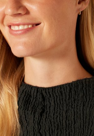 Close-up van een persoon die een zwarte top met textuur en een gerimpeld ontwerp draagt, met een kleine hoepeloorbel, en een gladde hals en huid toont.