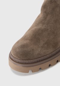 Bota de ante marrón hasta el tobillo con punta redonda y suela texturizada con surcos profundos para mayor tracción. El diseño es minimalista con una suela gruesa.