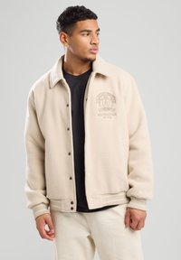 Beige fleece jacket with a collar, front buttons, and a logo on the left chest. Ribbed cuffs and hem with white stripes. Worn over a black shirt.