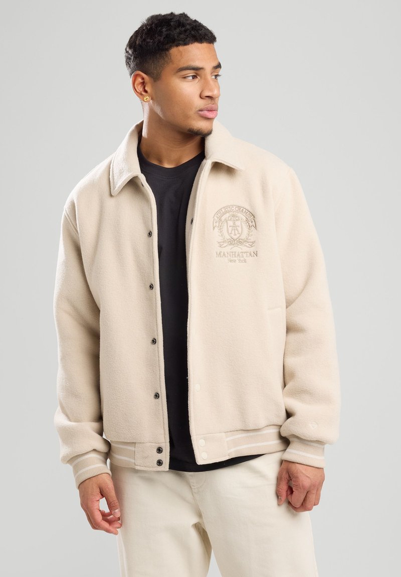 Beige fleece jacket with a collar, front buttons, and a logo on the left chest. Ribbed cuffs and hem with white stripes. Worn over a black shirt.