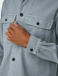 Camisa azul clara con dos bolsillos en el pecho, botones negros y puños abotonados. Tela suave y texturizada con un brillo sutil.