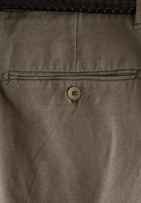 Pantaloni di tessuto beige con una tasca posteriore chiusa da un bottone marrone. La texture è liscia con un motivo sottile.