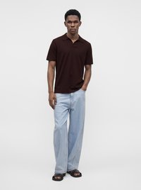 Jeune homme debout, une main dans la poche, vêtu d’un polo foncé, d’un jean ample bleu clair et de sandales noires sur un fond uni.