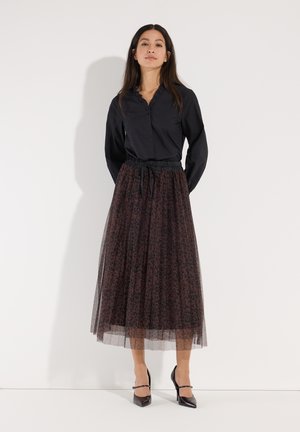 Vrouw staand tegen een witte achtergrond, gekleed in een zwarte blouse met lange mouwen, een bruine midi tulen rok met patroon en zwarte hakken.