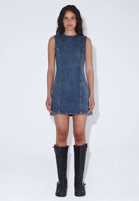 Robe sans manches en denim bleu foncé, présentant une silhouette ajustée et des coutures visibles. Portée avec des bottes noires montant jusqu'aux genoux.