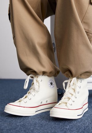White high-top Converse sneakers with platform soles worn with loose tan pants on blue carpeted floor.