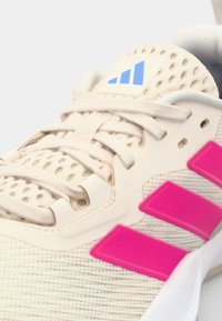 Lys beige sportsko med rosa striper; har et pustende mesh-overdel, snøringssystem og teksturert såle. Blå logo på tungen.