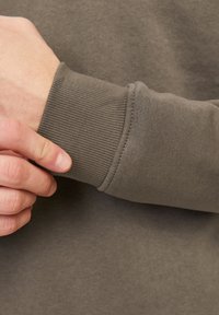 Close-up van bruine sweatshirtstof die een ribbelmanchet met stikdetails toont, naast een gedeeltelijk zichtbare hand. Over het algemeen een gladde textuur.