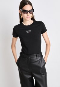 Μαύρη κοντή μπλούζα εφαρμοστού τύπου με το λογότυπο "tommy jeans", συνδυασμένη με ψηλόμεσα μαύρα δερμάτινα παντελόνια. Το μοντέλο φοράει υπερμεγέθη μαύρα γυαλιά ηλίου και μια κολιέ από μαργαρίτες.