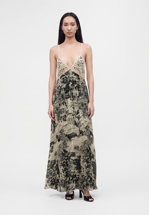 Femme aux longs cheveux foncés portant une robe longue sans manches à encolure en V avec un haut en dentelle et un imprimé nature noir et beige, debout, les bras détendus.