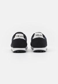 Crne zamšaste tenisice s bijelim gumenim potplatima, koje imaju podstavljenu ovratnik i istaknuti "VEJA" logo na peti. Texturirani detalji na zadnjem dijelu.