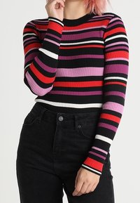 Persona che indossa un maglione a costine con maniche lunghe e righe orizzontali in nero, rosso, viola, rosa e bianco, abbinato a pantaloni neri a vita alta.