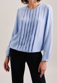Blusa azzurro chiaro a maniche lunghe con pieghe verticali, scollatura rotonda e texture liscia, abbinata a pantaloni scuri.